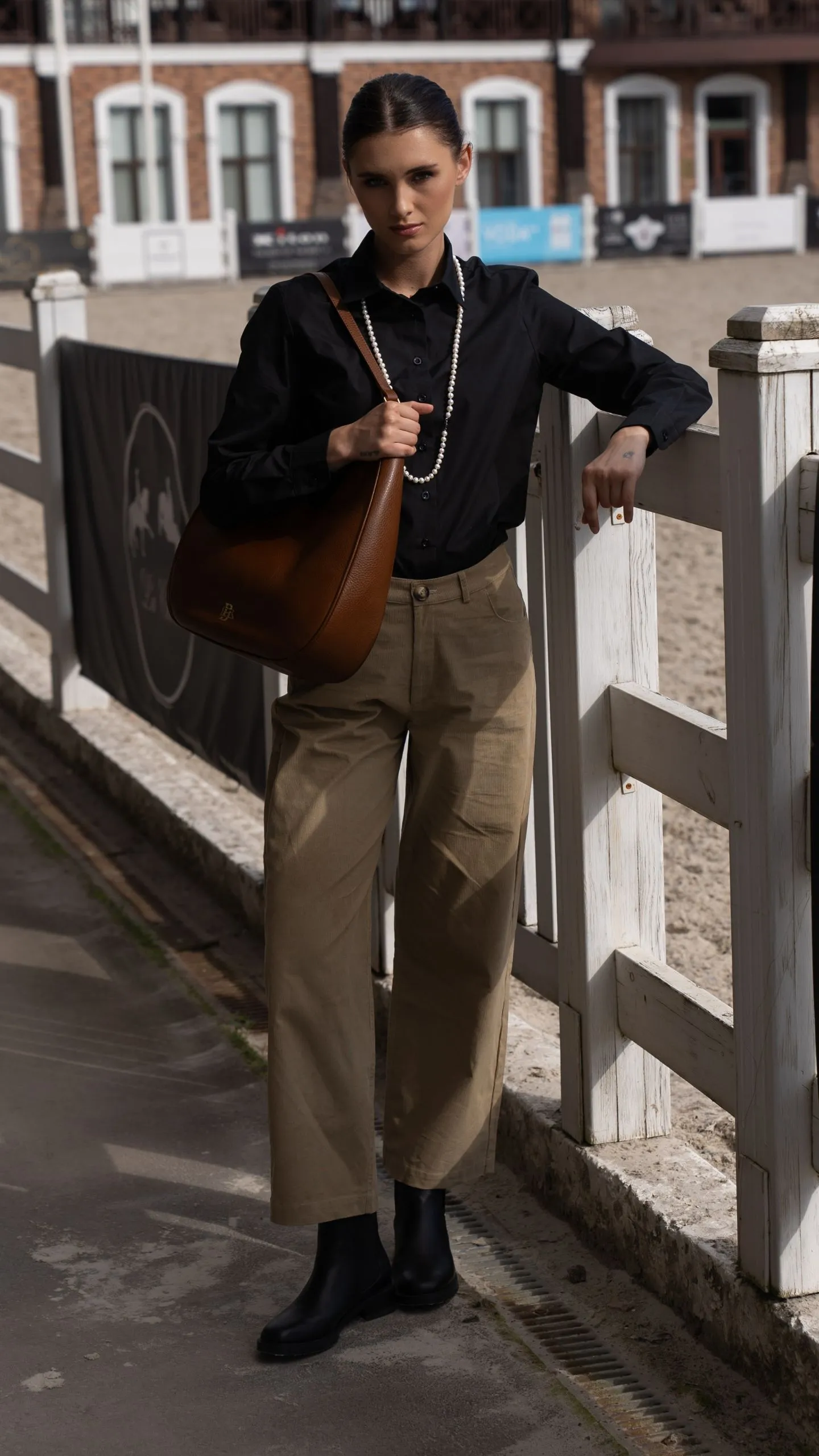 Дівчина в чорній сорочці, бежевих прямих штанах і з коричневою сумкою через плече 